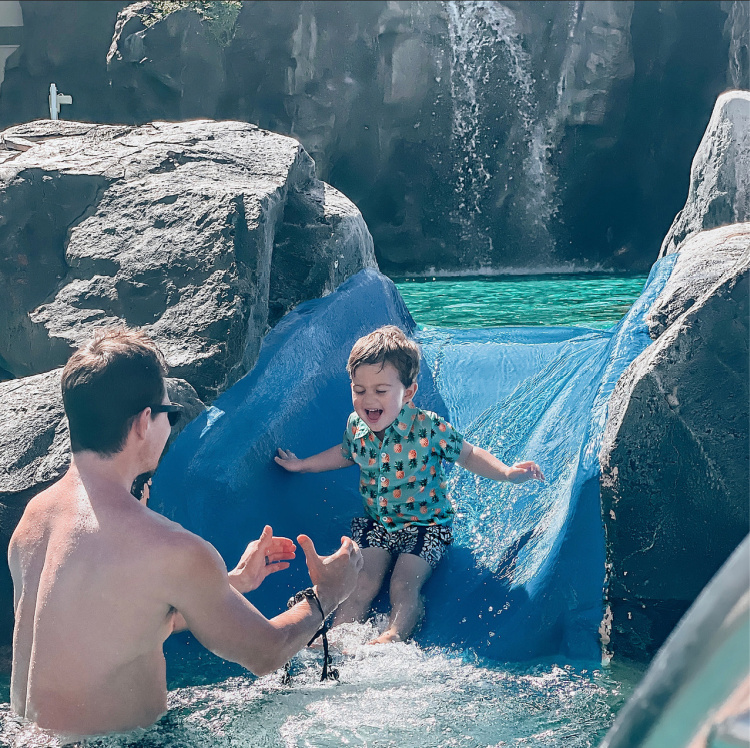 Jake sliding down the water slide in the resort pool being caught by Kyle