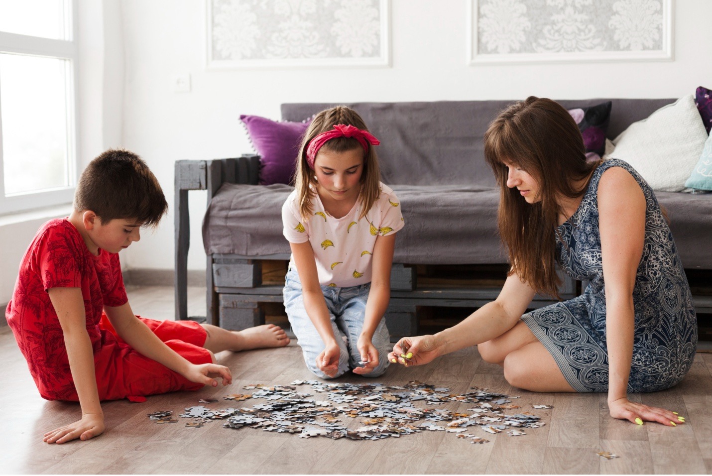 Two kids and a woman sitting on the floor in front of a couch with puzzle pieces spilled out in front of them.