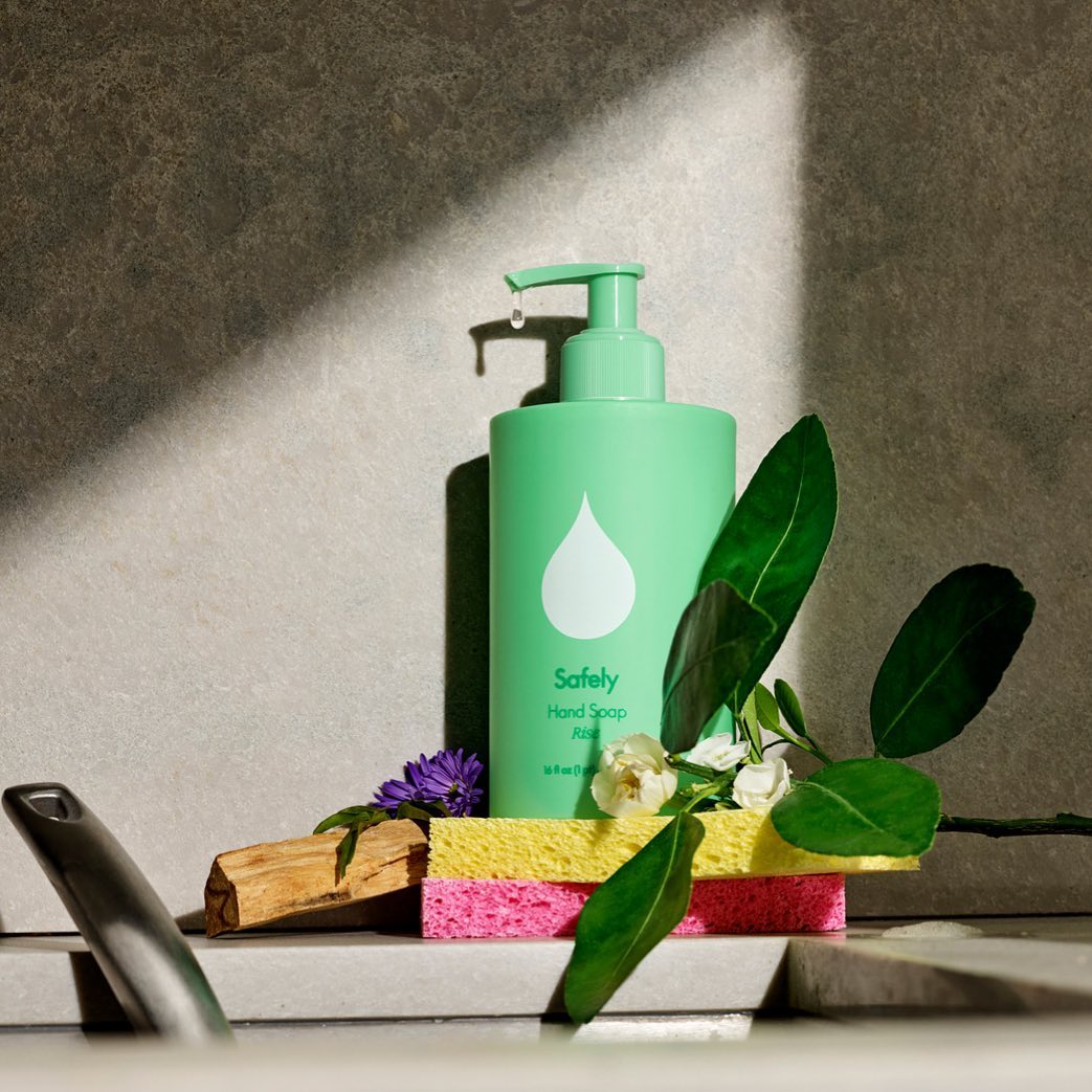 Bottle of green Safely hand soap sitting behind sponges and green leaves by a sink.