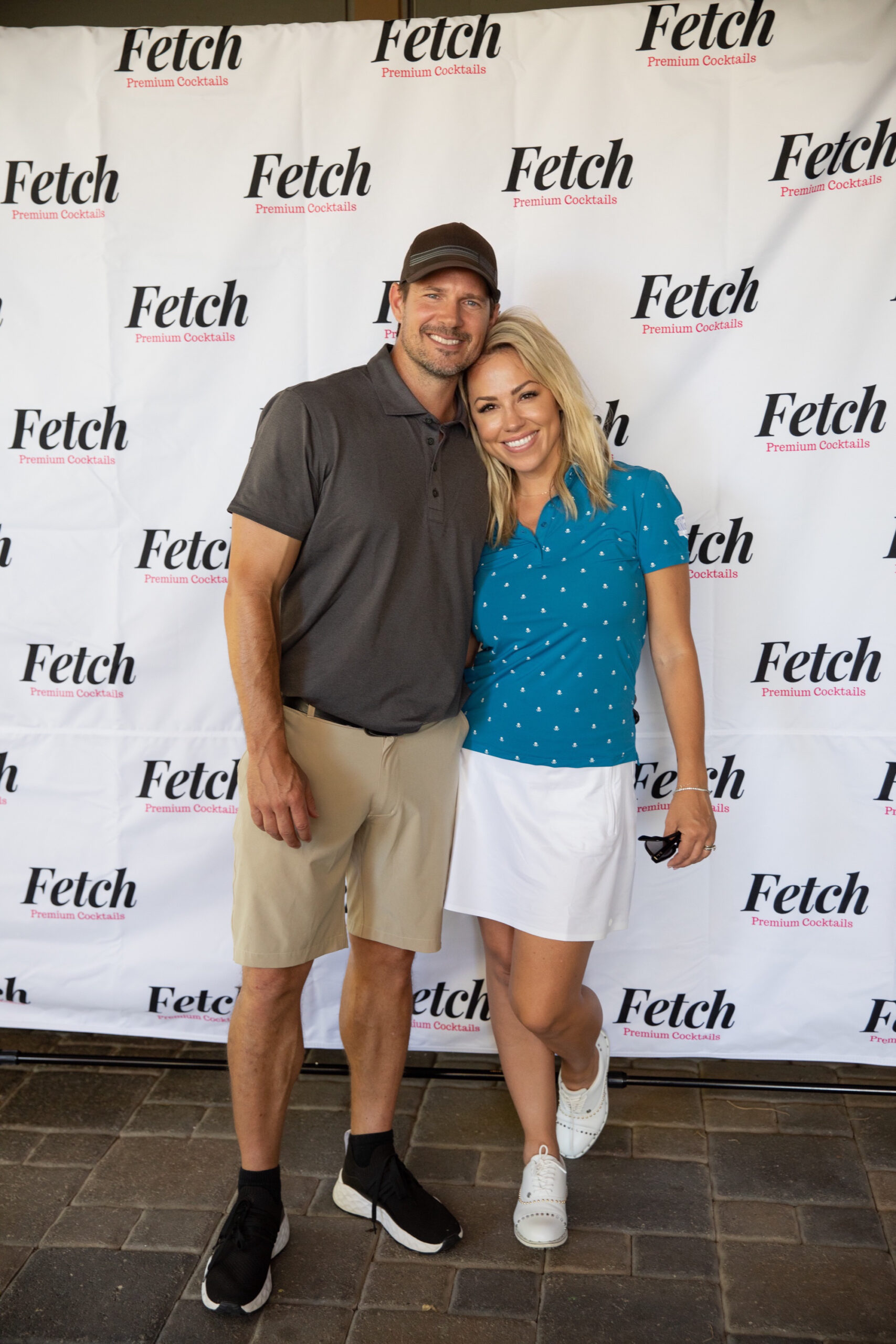 Jessica Hall and husband, Kyle, wearing golf clothes and standing in front of a step and repeat with Fetch Cocktail logos.