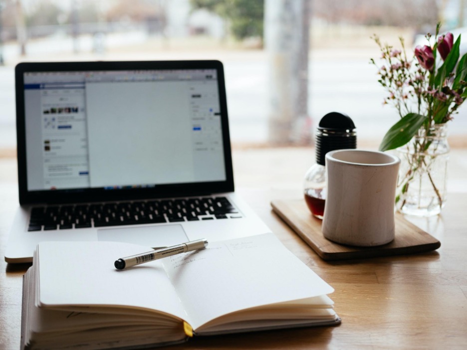 Laptop computer, coffee mug, and journal with pen sitting on a desk in front of a window.