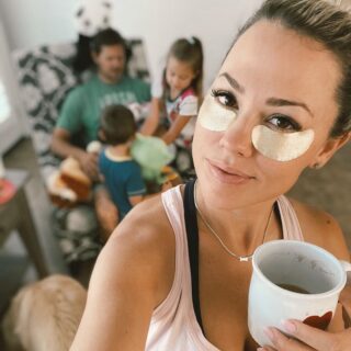 Jessica Hall holding a coffee cup and wearing gold under-eye strips with her family blurred in the background.