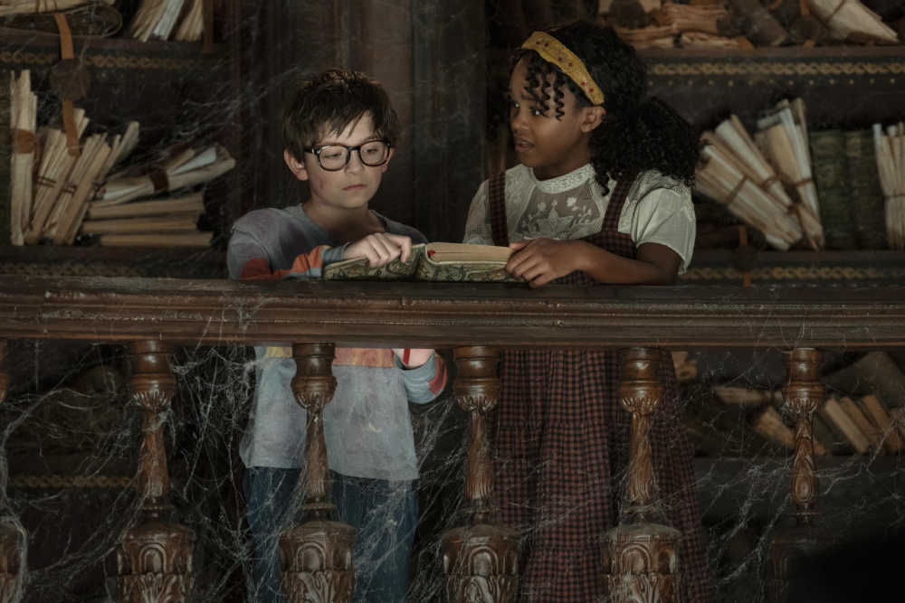 Winslow Fegley and Lidya Jewett from the movie NIghtbooks standing in a dirty library looking at a book.